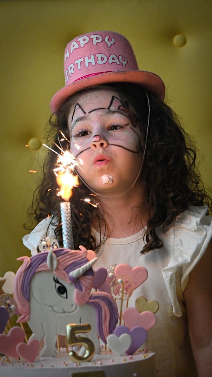 A young girl celebrates her birthday with a unicorn cake and sparklers indoors.
