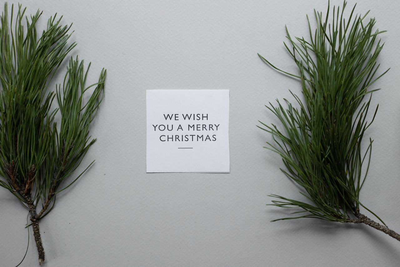Top view composition of We Wish You A Merry Christmas inscription on white paper sheet on table with spruce twigs on gray background